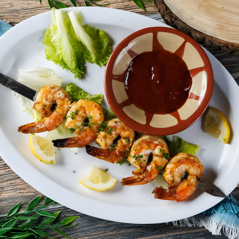 Grilled shrimp kabob with lemon wedges, lettuce, and a side of red sauce on a white plate.